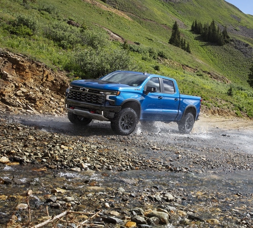 A blue pickup truck drives through a rocky stream against a backdrop of lush green hills.