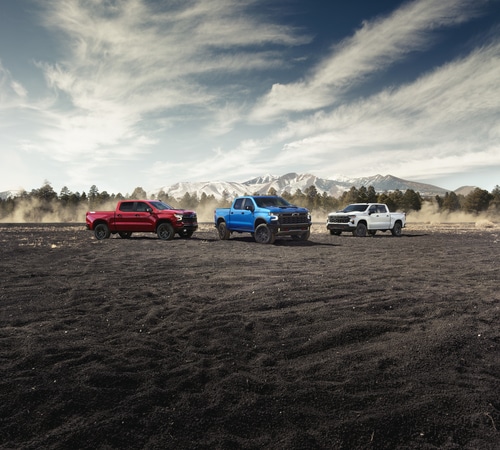 Three trucks in red, blue, and white parked on rugged terrain with a mountainous backdrop under a partly cloudy sky.
