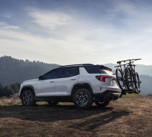 A white SUV with two bicycles mounted on the back parked on a grassy hill, scenic forest view.