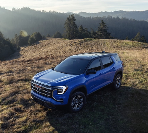 Blue SUV parked on a grassy hilltop, surrounded by scenic forested mountains.