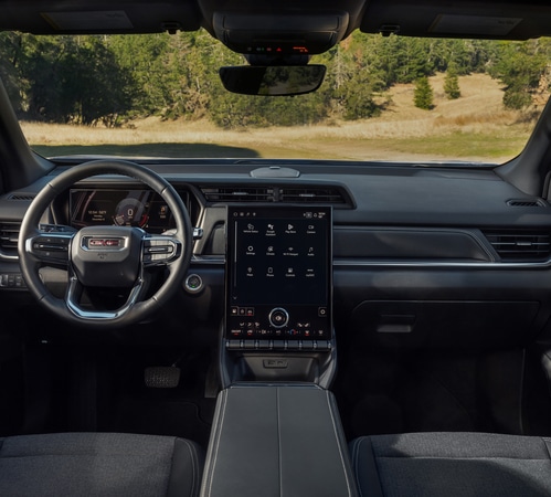 Interior view of a modern car dashboard featuring a large touchscreen and steering wheel. Forest scenery outside.