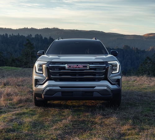 Front view of a silver GMC SUV parked in a grassy field with mountains in the background.