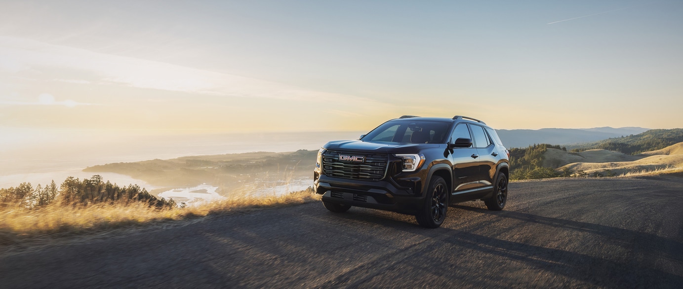 A green SUV is parked on a paved surface near a calm body of water. The car features a prominent grille and sleek design. In the background, hills are visible under a clear blue sky.