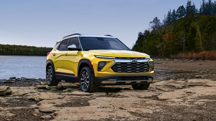 Yellow SUV parked on rocky shore by a lake, surrounded by trees under a clear sky.