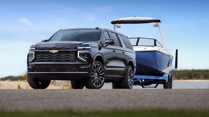 Chevrolet SUV towing a blue boat on a road near a lake, under a clear blue sky.