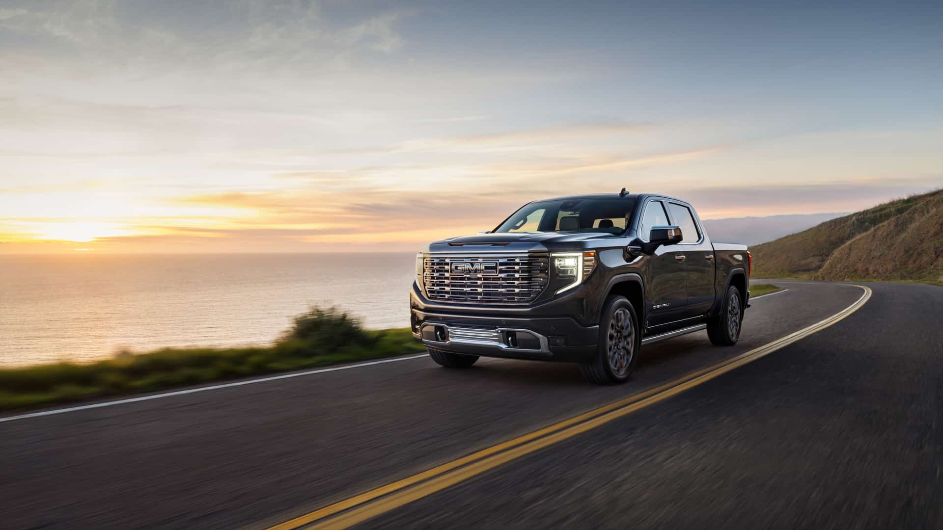 GMC truck driving on a coastal road at sunset, with ocean and cliffs in the background.