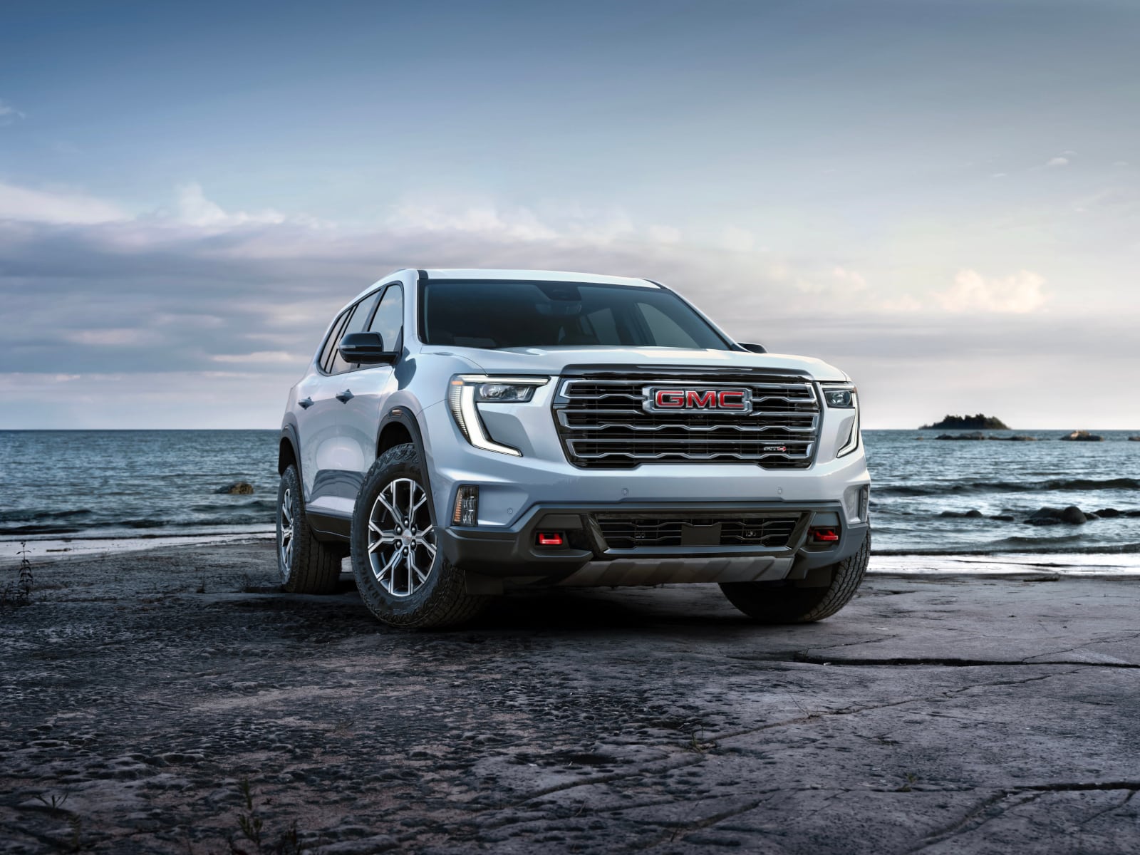 Silver GMC SUV parked by the ocean on a rocky beach under a cloudy sky.