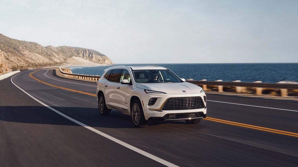 White SUV driving on a coastal highway with scenic ocean and mountain views.